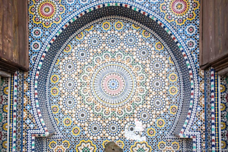 Close-up of a wall fountain covered in dense multicolored geometric mosaic arranged around a circular rosette.