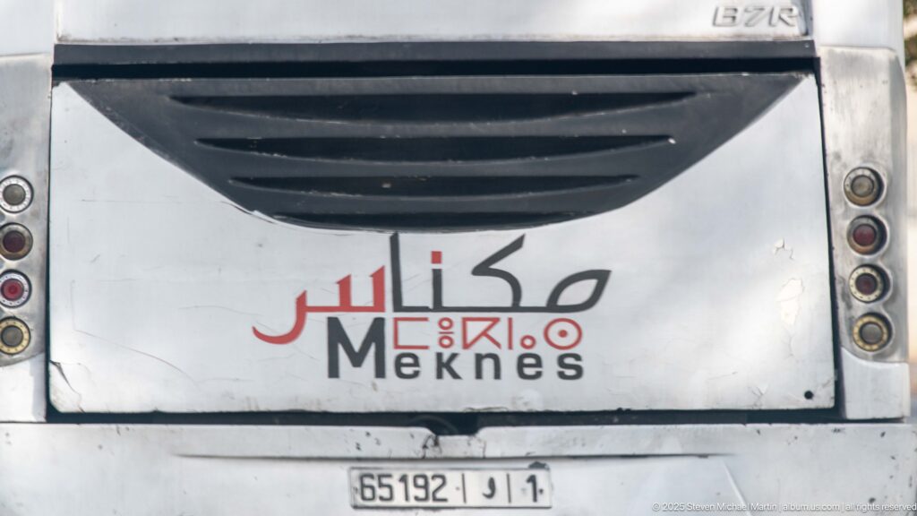 Close rear view of a white coach marked Meknes in Arabic and Latin lettering.