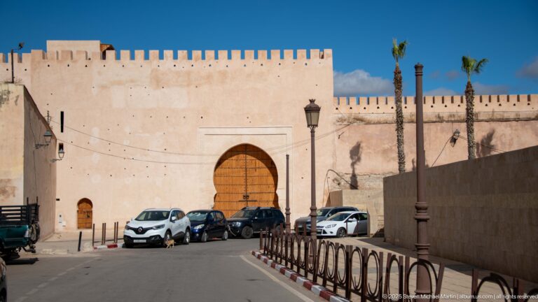 Plain wooden arched doorway set in a broad crenellated wall, with parked cars along the street.