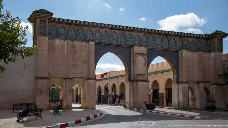 Two tall horseshoe arches span a roadway beneath a blue-green decorative frieze, with shops and arcades beside them.