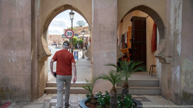 Man walking through shaded horseshoe arches toward a sunlit street, beside a small planted bed and shop doorway.