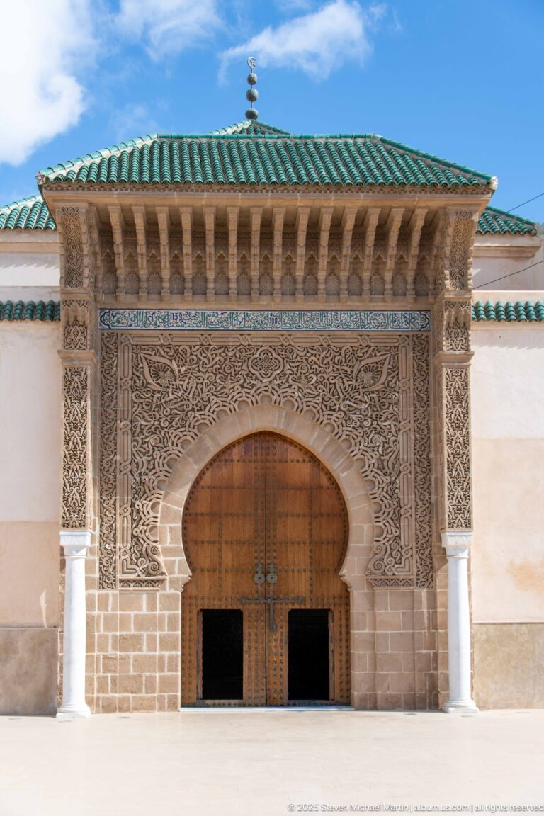 Large arched wooden door surrounded by dense carved ornament beneath a projecting green-tiled roof.