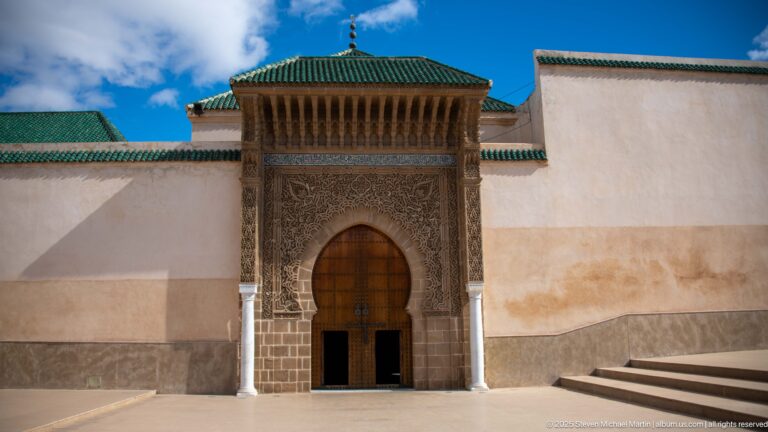 Tall carved portal with wooden door and green-tiled canopy centered in a broad pale wall.