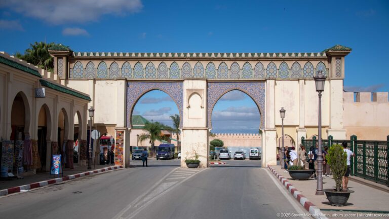 Street perspective toward a twin-arched gateway with blue-green frieze, shops, planters, pedestrians, and cars.