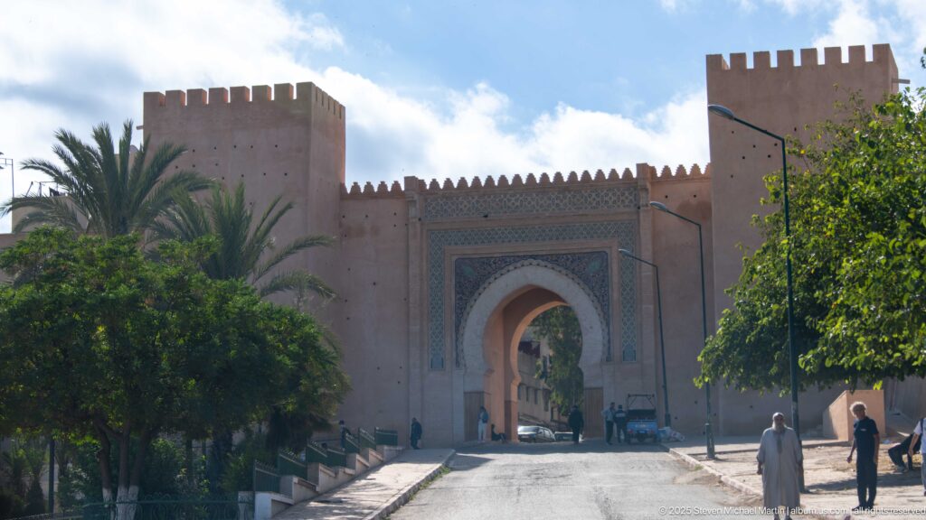 Decorated horseshoe-arched gate between crenellated towers, with palms, pedestrians, and a road passing through.