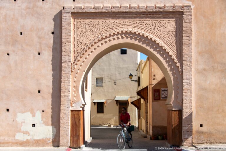 Carved horseshoe-arched gateway in an earthen wall framing a cyclist on a narrow street.