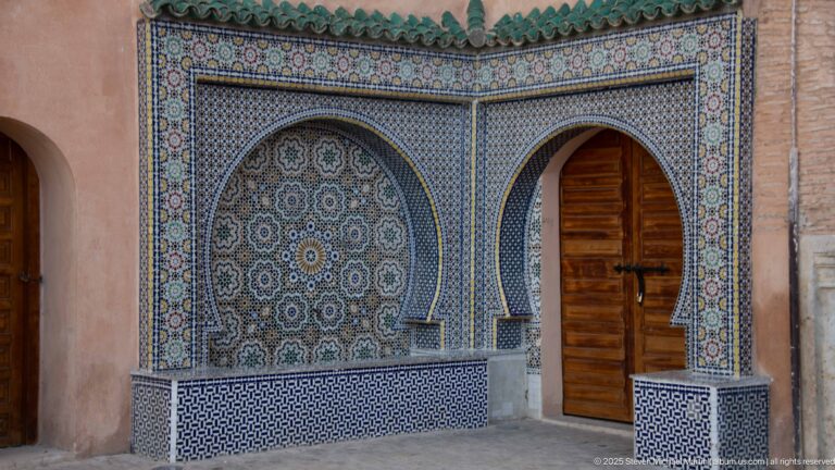 Zellij-covered wall fountain with circular geometric motifs beside a tall wooden arched doorway.