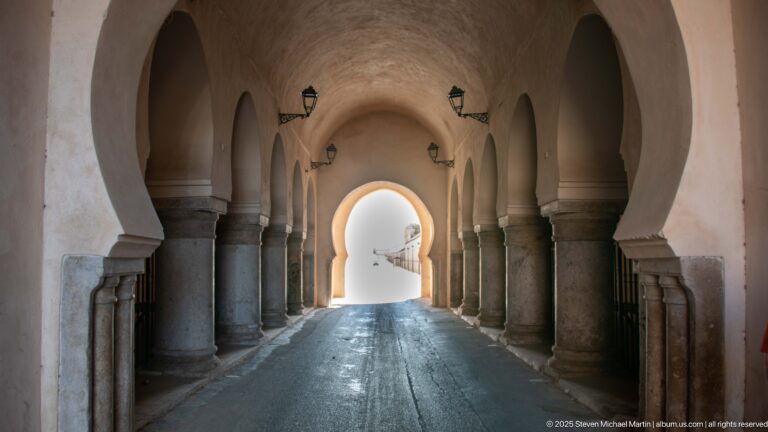 Long vaulted passage with repeated horseshoe arches, stone columns, wall lanterns, and bright daylight at the far end.