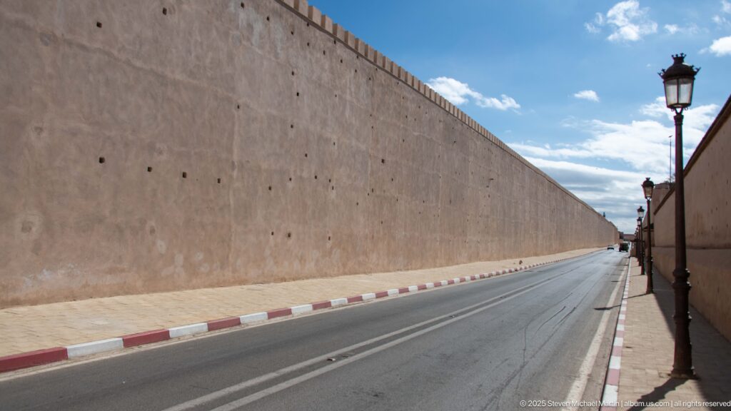 Long pale fortification wall beside an empty paved road with red-and-white curbs and street lamps.