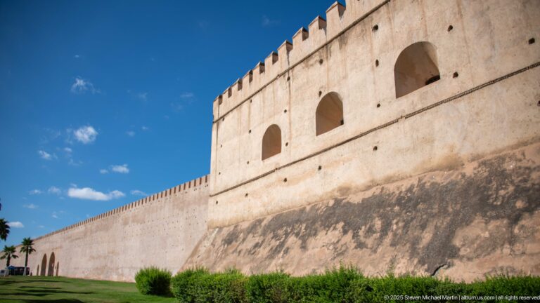 Fortified wall with a projecting block, large arched openings, crenellated parapet, and sloped weathered base.