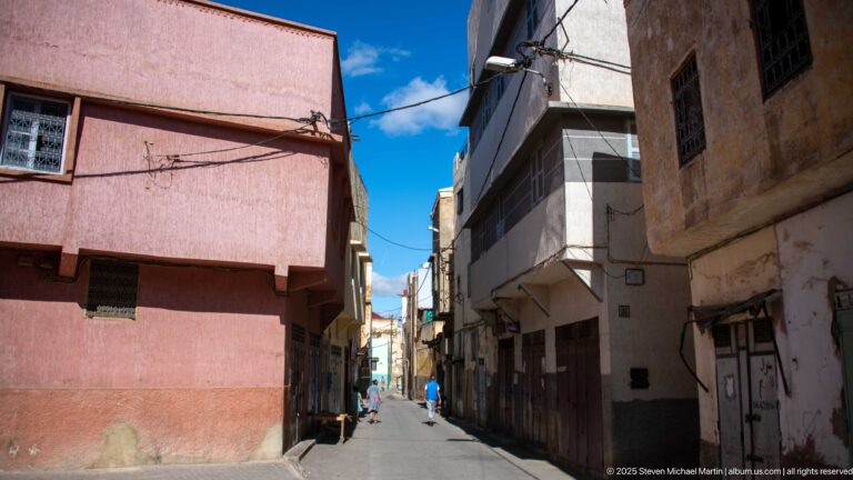 Narrow urban street with pink and weathered façades, overhead wires, doors, windows, and distant pedestrians.