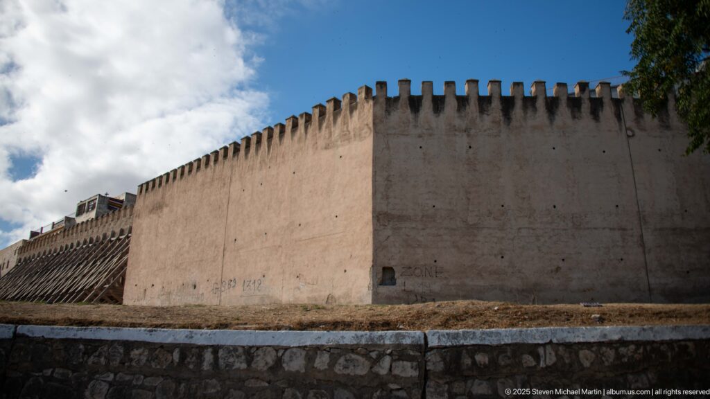 Long pale earthen fortification wall with a crenellated parapet rising above a stone base.