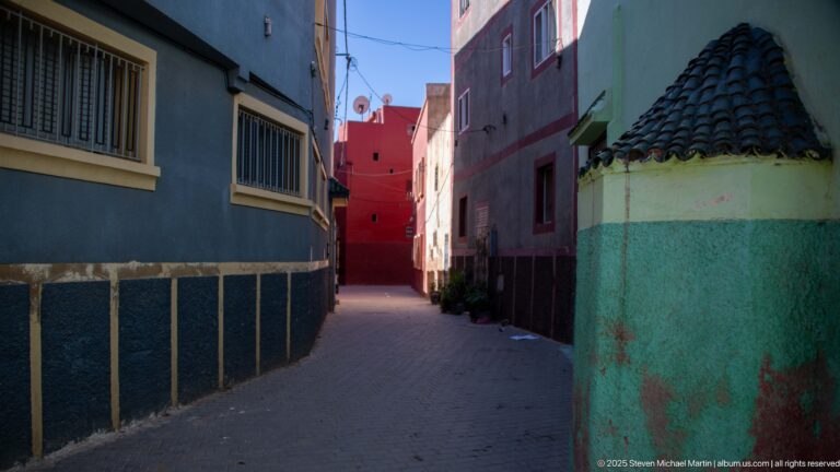 Narrow pedestrian lane between blue, green, purple, and red houses with barred windows and a small tiled roof.