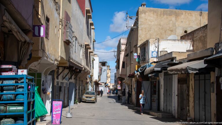 Narrow Meknes street with weathered buildings, shops, pedestrians, parked car, overhead wires, and stacked gas cylinders.