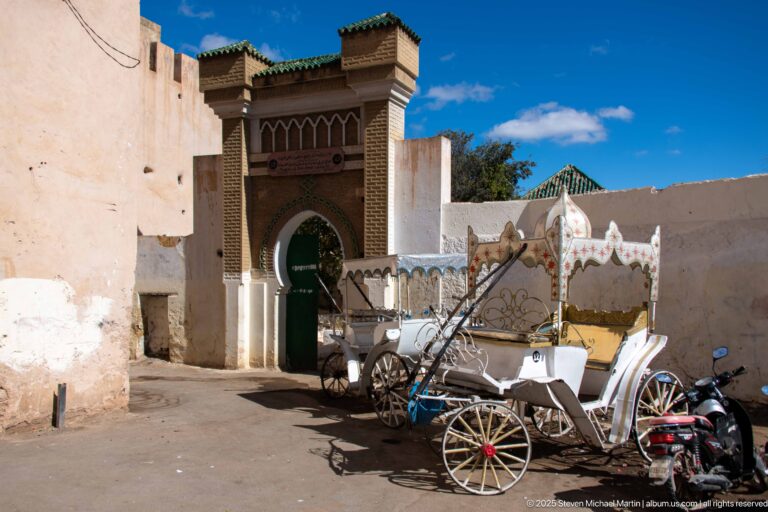 White decorative carriage parked before a carved portal with a green arched door and tiled roof.