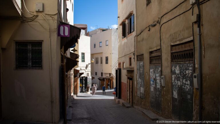 Narrow shaded lane between weathered buildings leading toward pedestrians in a brighter street beyond.