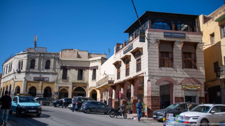 Busy Meknes street corner with cars, motorbikes, pedestrians, arcaded shops, and mixed-height buildings.
