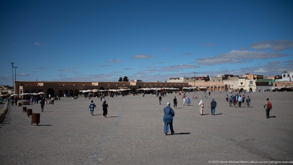 Large paved square with scattered pedestrians and denser market activity along the far side.