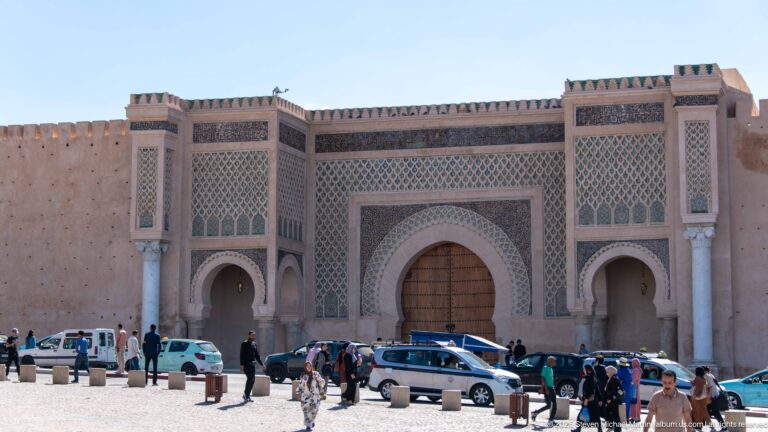 Large decorated gateway with a central arched wooden door, smaller side arches, patterned panels, crenellations, people, and cars.