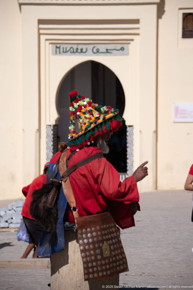 Moroccan water seller in red clothing and a colorful tasselled hat carrying a leather water bag and studded pouch.