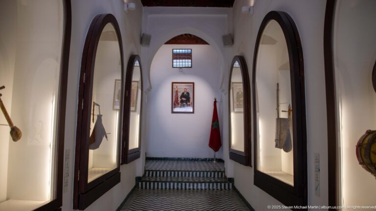 Long museum corridor with arched display recesses, striped tile floor, official portrait, and Moroccan flag at the far end.