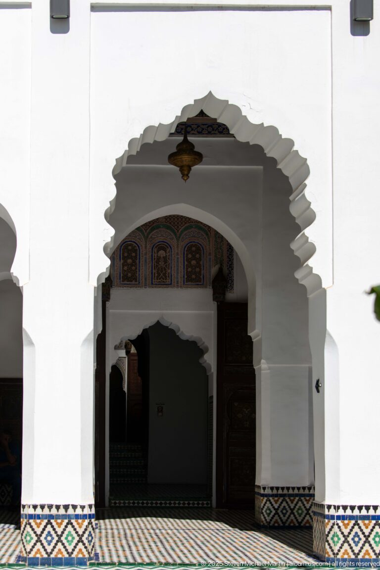 White museum interior with a scalloped arch, low geometric zellij, hanging lantern, and darker carved doorway beyond.
