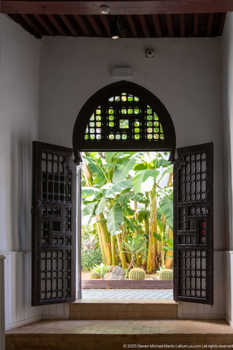 Open dark wooden lattice doors beneath an arched transom framing a lush green courtyard garden.