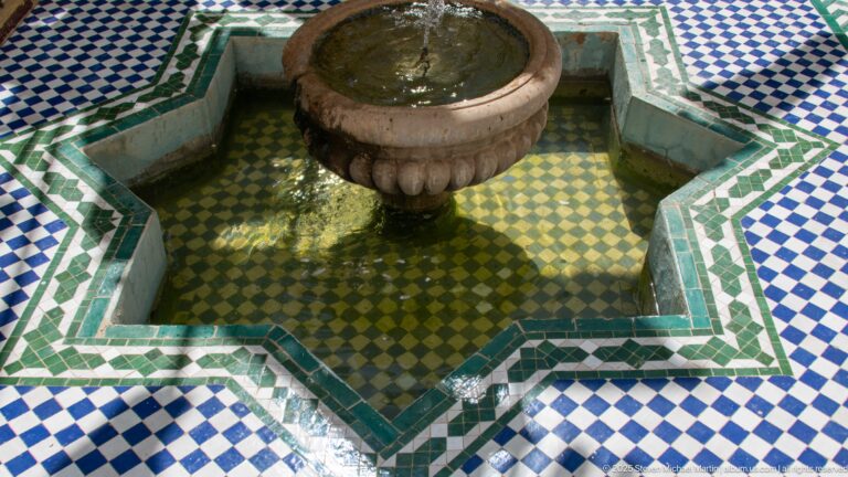 Stone fountain centered in a star-shaped zellij basin with green, blue, and white geometric tile patterns.