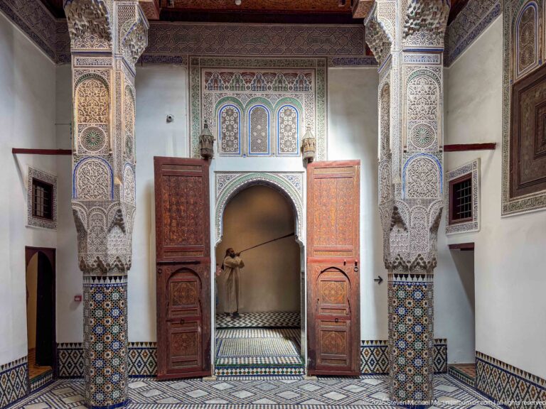 Open interior doorway surrounded by carved ornament, zellij, painted wood panels, and slender decorated columns.