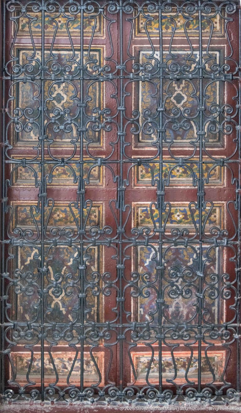 Close-up of black wrought-iron scrollwork grille covering reddish carved wooden double doors.