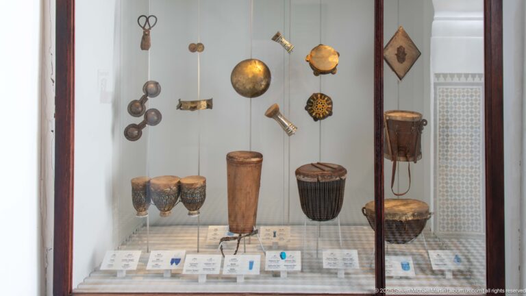 Museum case with tall drums, small hand drums, cymbals, and suspended metal percussion instruments.