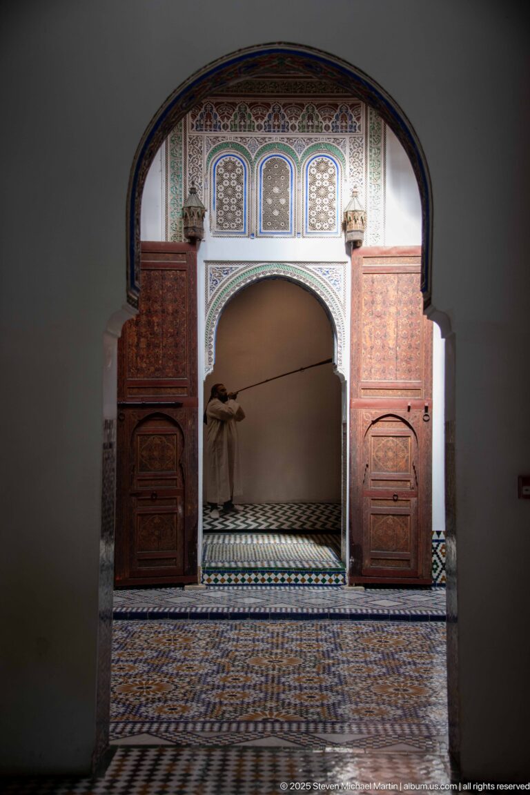 Dark foreground arch framing an ornate room with painted wood panels, a scalloped opening, and patterned tile floor.