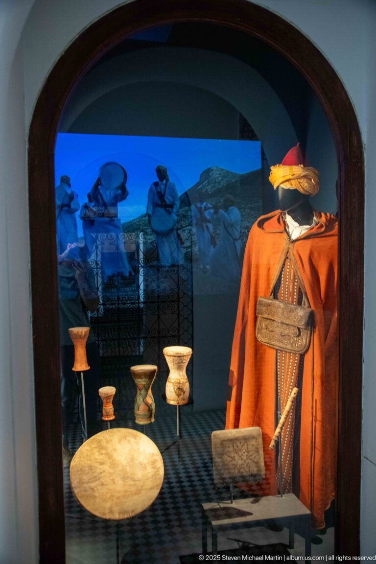 Red traditional garment with hat, belt or pouch, and small metal vessels displayed before a blue backdrop.