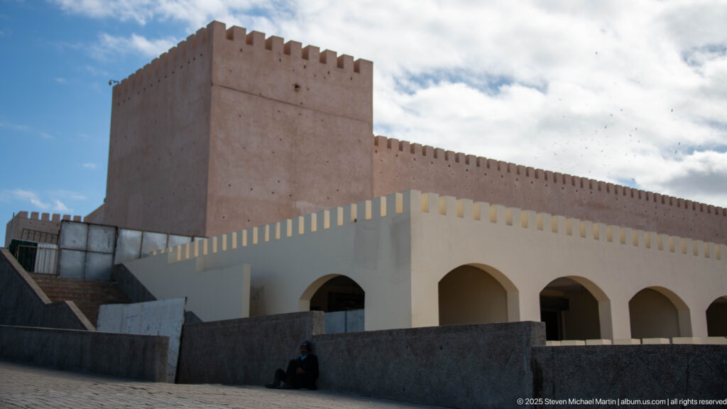 Square crenellated tower and wall rise behind a lower white building with rounded arches.