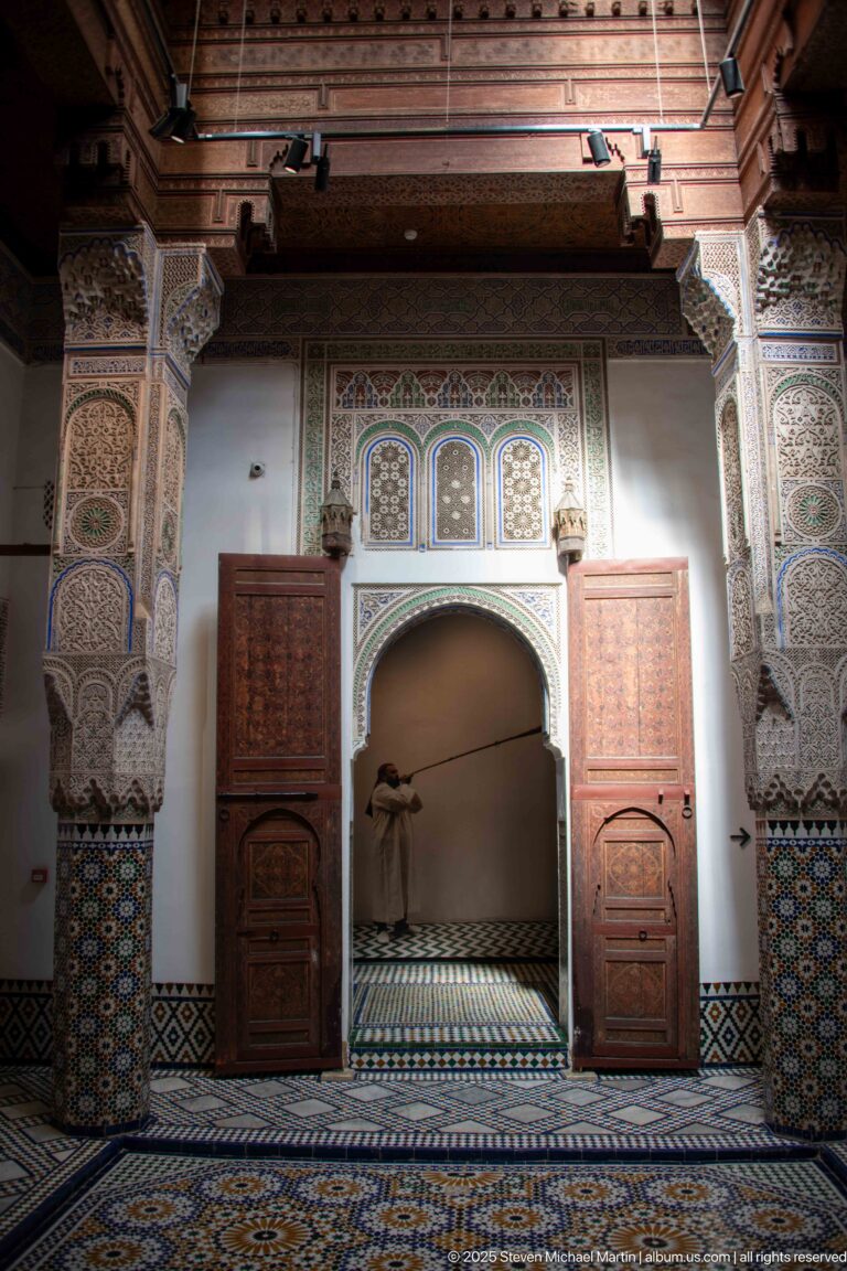 Ornate room with scalloped arch, painted wooden panels, carved supports, lanterns, and patterned tile floor.