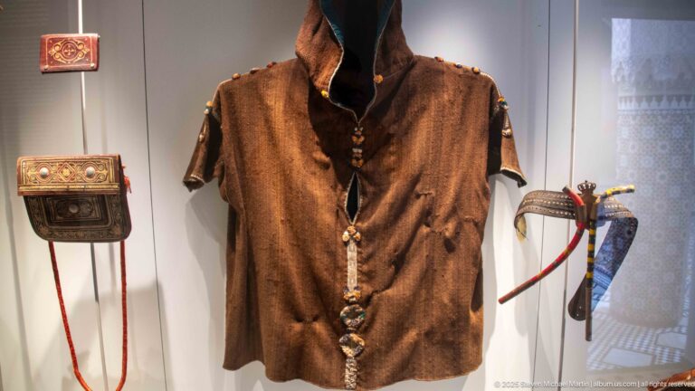 Brown sleeveless garment displayed with a decorated pouch and sheathed curved blade or dagger.