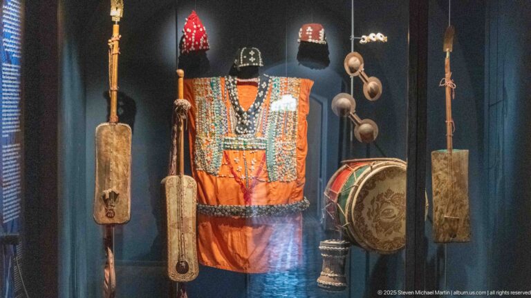 Orange and turquoise costume displayed with drums, cymbals, rattles, and long traditional instruments.