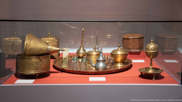 Museum case with engraved metal ewers, covered vessels, bowls, and perforated containers on a red plinth.