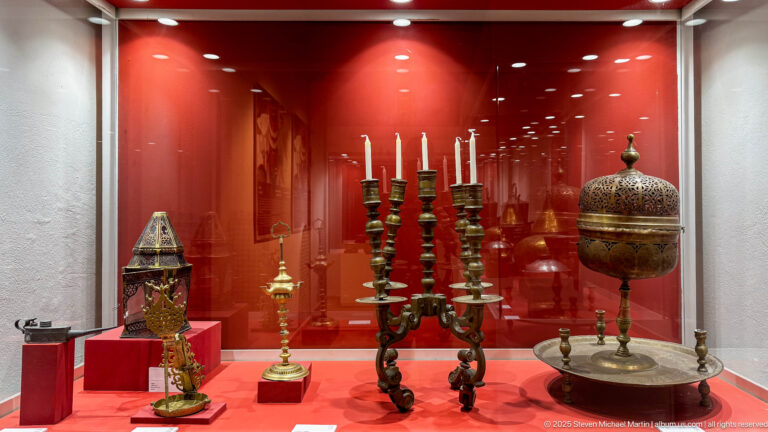 Museum case with tall candlesticks, pierced lanterns, and covered metal lighting objects against a red background.