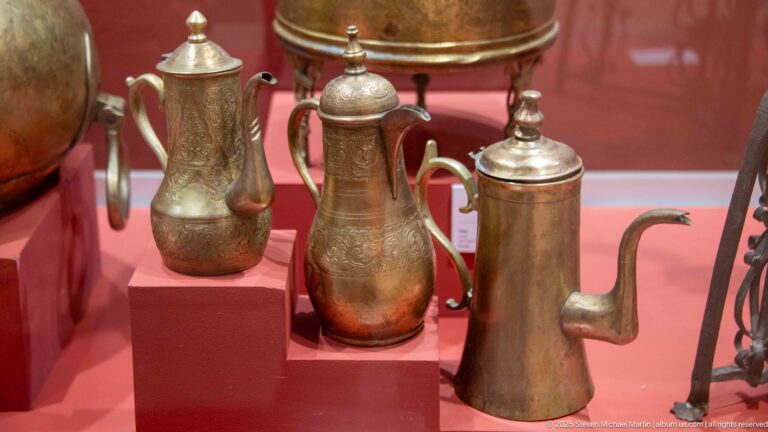 Three engraved metal ewers with curved spouts, lids, and handles displayed side by side.
