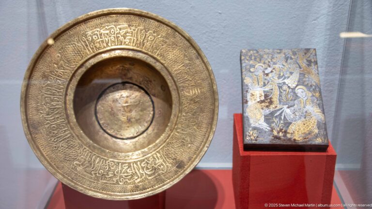 Large engraved brass-colored basin with Arabic calligraphy around its rim beside a smaller decorated object.