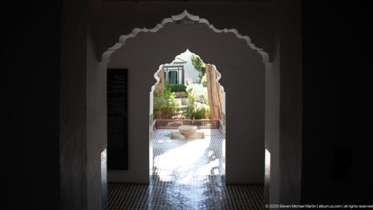 Dark scalloped arch framing a bright planted courtyard with tiled paving and a central fountain or basin.