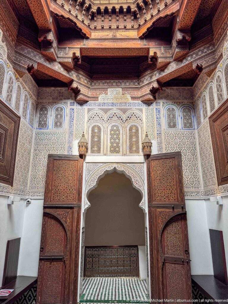 Ornate interior wall with scalloped arch, carved ornament, painted wooden doors, brackets, and decorated ceiling.