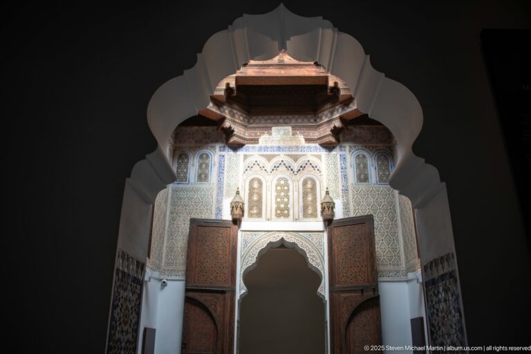 Tall scalloped arch framed by carved plaster, zellij, painted wood panels, and a projecting wooden ceiling.
