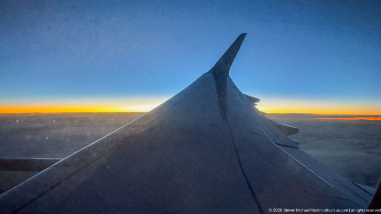 Airplane wing above dark clouds with a thin orange glow along the horizon under a deep blue sky.