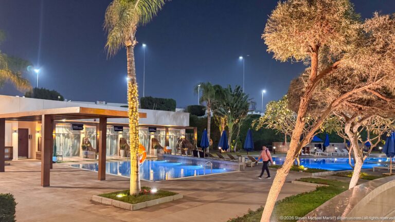 Illuminated hotel swimming pool at night with palm trees, lounge chairs, blue umbrellas, and low buildings.