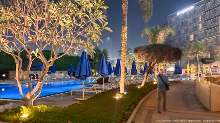 Curved walkway beside a blue-lit hotel pool with illuminated palms, lounge chairs, umbrellas, and hotel building.