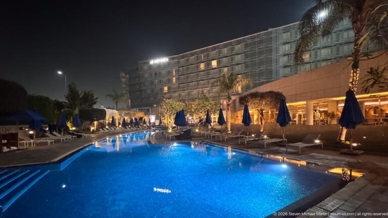 Large blue-lit swimming pool at night with closed umbrellas, empty lounge chairs, palms, and hotel façade.