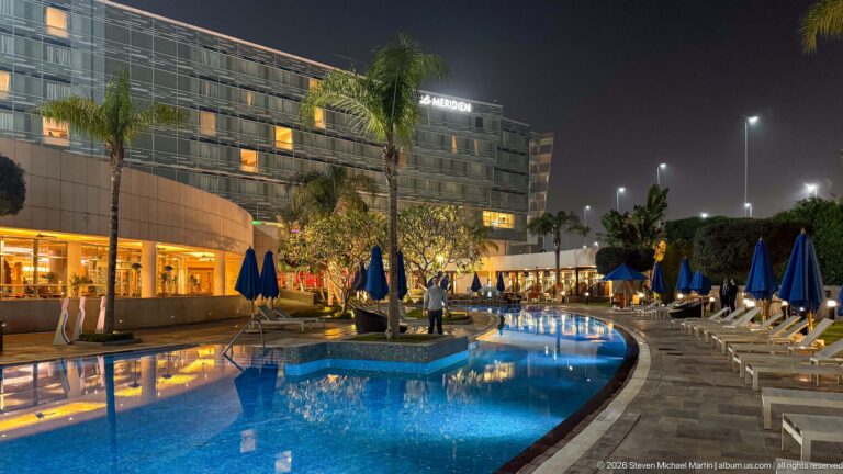 Curving illuminated hotel pool with palm trees, blue umbrellas, lounge chairs, and a large building at night.