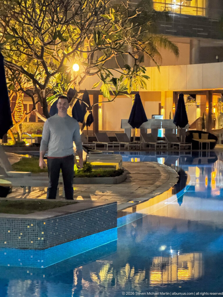 Man standing on a tiled platform beside a glowing hotel pool, with trees, loungers, and umbrellas at night.
